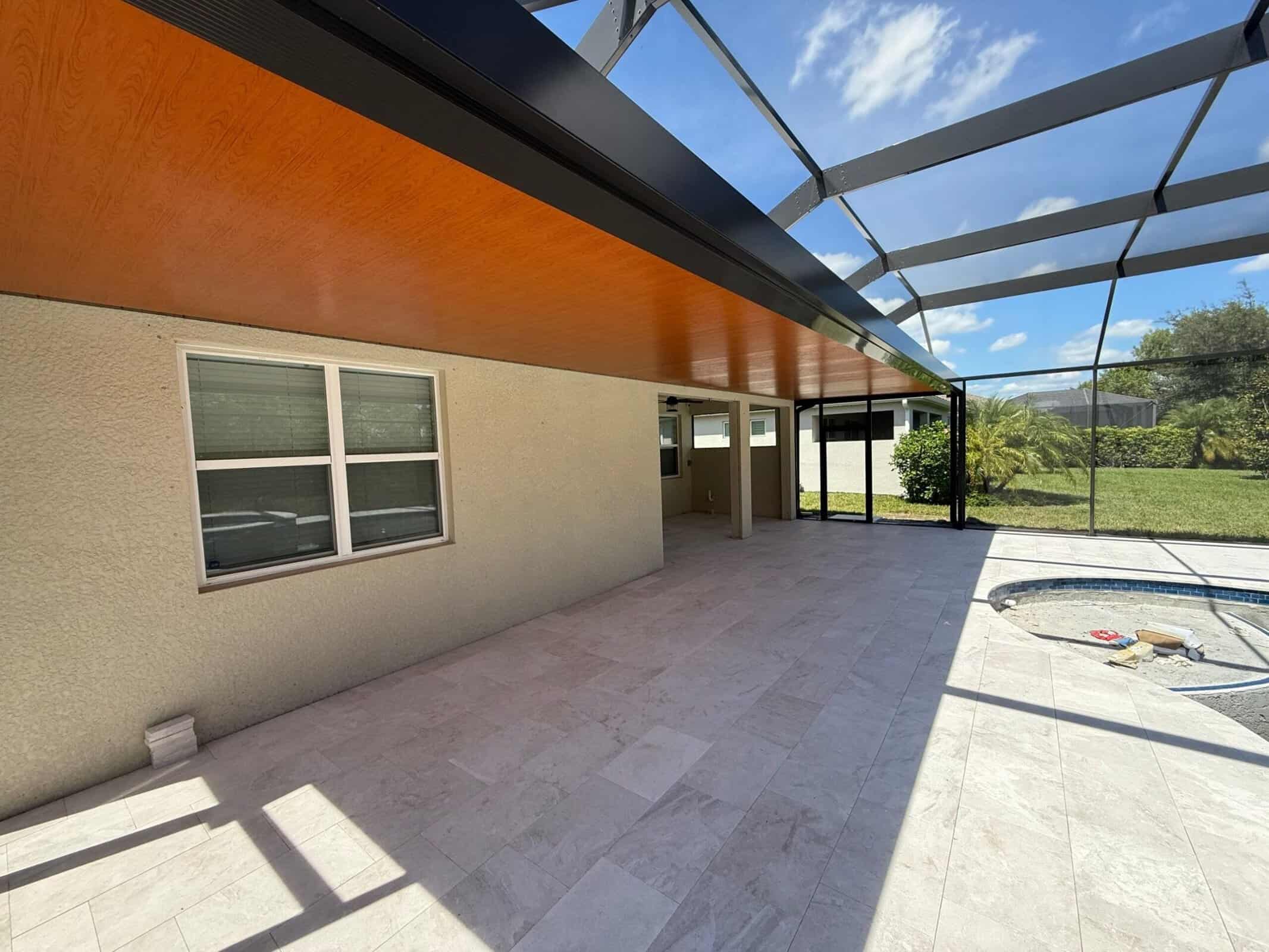 Wood-look covered patio ceiling meeting dark aluminum pool cage framing at Stillwater in Naples, with the pool still under construction.